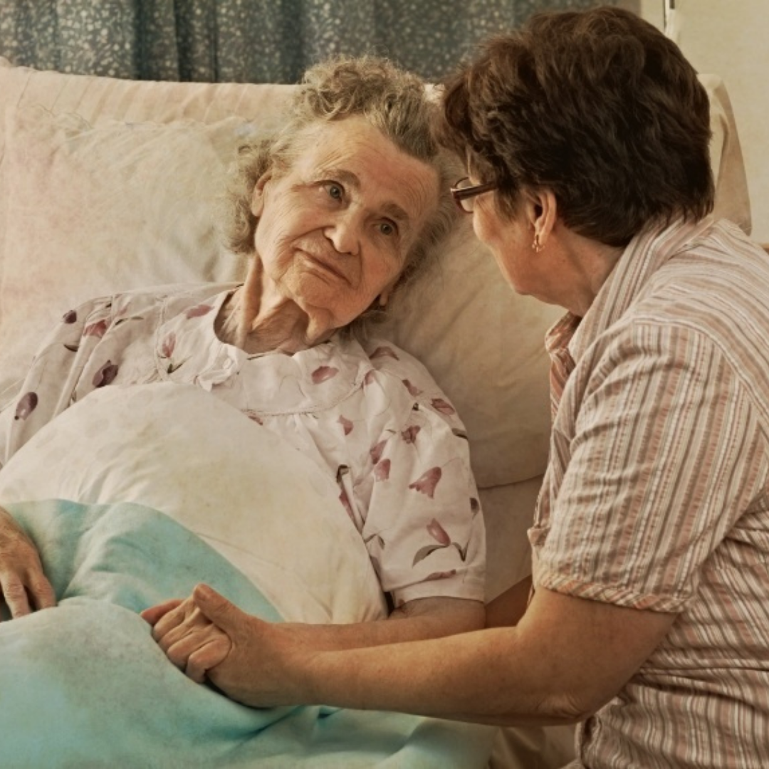 an elderly lady in a hospital bed and a companion right beside her holding her hand